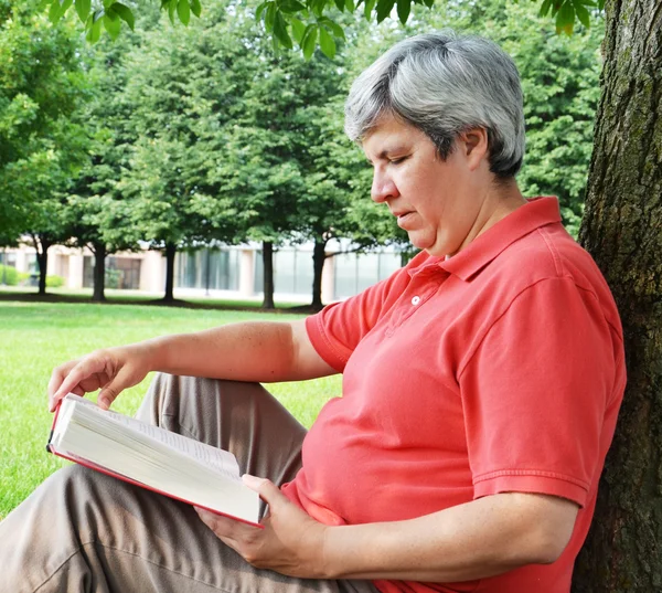 Mulher de meia-idade lendo livro por árvore — Fotografia de Stock