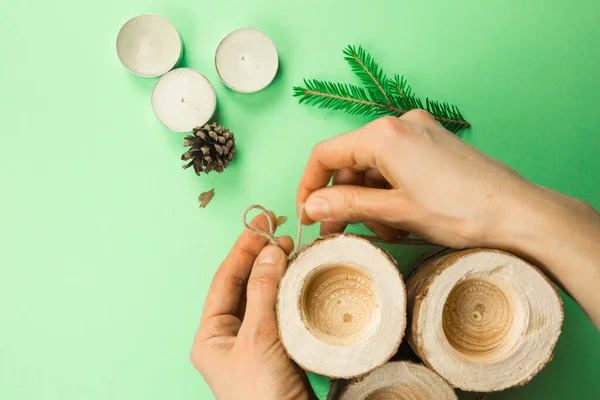 DIY Christmas candle holder made of pine logs, candles, craft rope, fir branches and cones. Hands fasten and tie the rope. Step-by-step instructions flat lay, step 3.