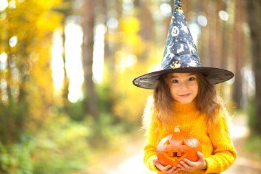 Sarı kazaklı, gözleri oyulmuş bir balkabağı ve sarı bir sonbahar ormanında ağzı olan cadı şapkalı bir kız. Cadılar Bayramı Tatili