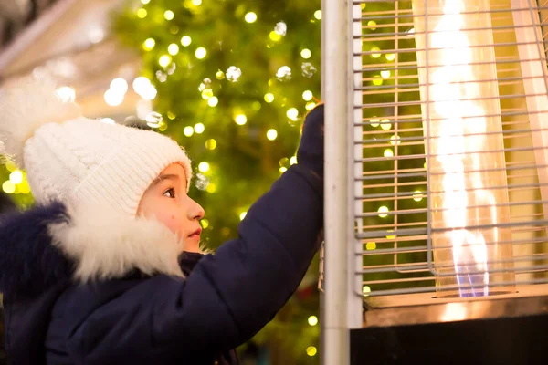 Girl in a warm hat looks at the flame of a gas burner warming at a Christmas market in the city, warms your hands. lights of garlands in defocus on the christmas tree, new year, festive festivities