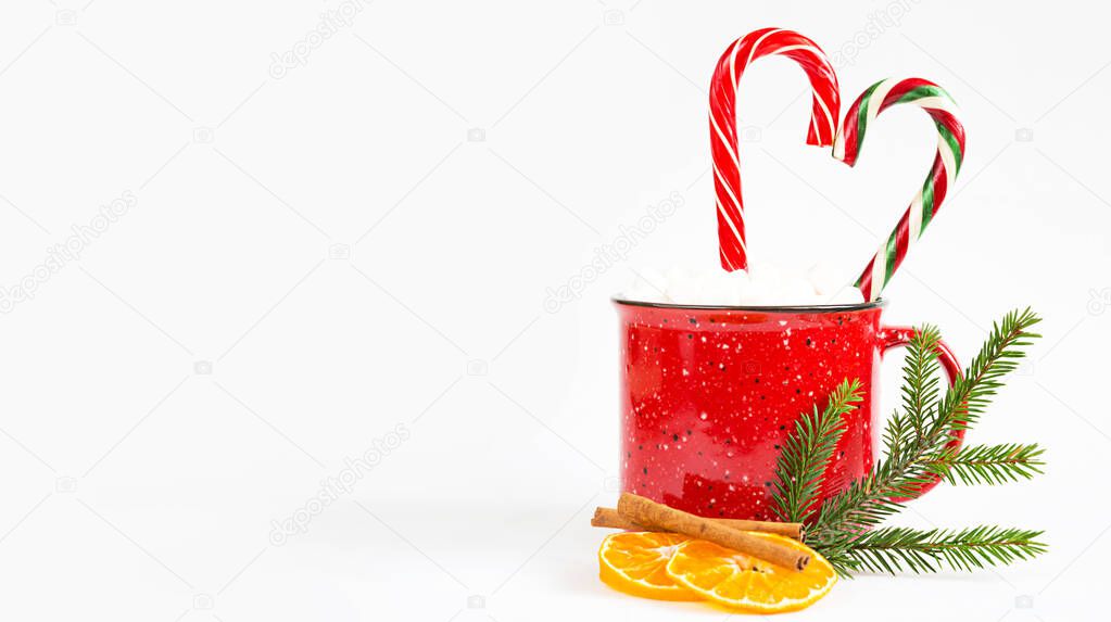 A red mug with marshmallows and candy cane in the shape of a heart is decorated with a natural fir branch or dried orange slices and cinnamon sticks. Christmas and New Year, the flavor of the holiday