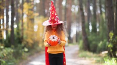 Sarı kazaklı, gözleri oyulmuş bir balkabağı ve sarı bir sonbahar ormanında ağzı olan cadı şapkalı bir kız. Cadılar Bayramı Tatili