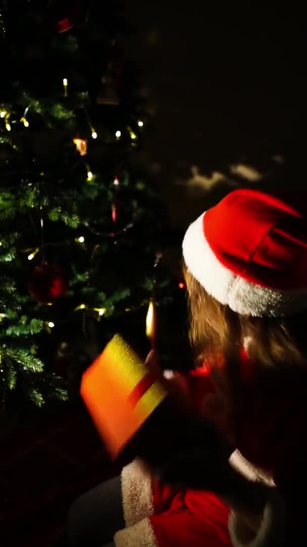 クリスマスツリーの近くにサンタクロースの衣装を着た女の子が夜にギフトボックスを開きます クリスマスの夜 子供の感情 奇跡の期待 垂直ビデオ — ストック動画