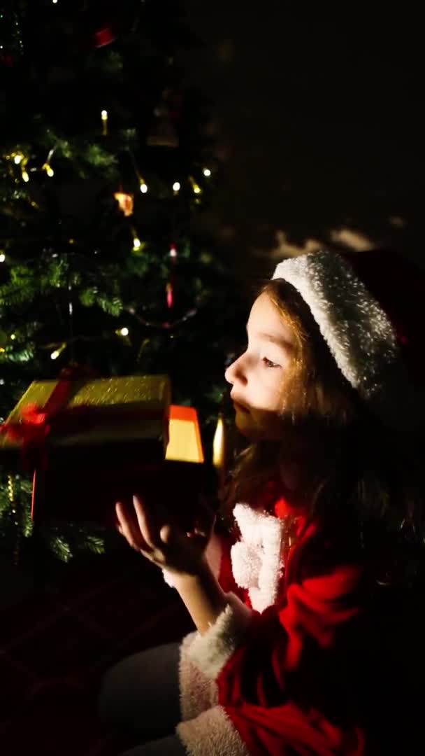 一个穿着圣诞老人服装的女孩在圣诞树旁 晚上打开礼品盒 平安夜 孩子们的感情 对奇迹的期待 垂直录像 — 图库视频影像