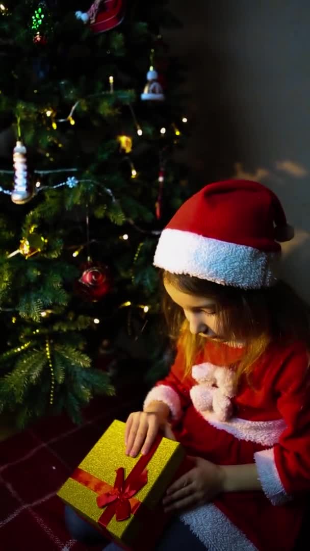 一个穿着圣诞老人服装的女孩在圣诞树旁 晚上打开礼品盒 平安夜 孩子们的感情 对奇迹的期待 垂直录像 — 图库视频影像