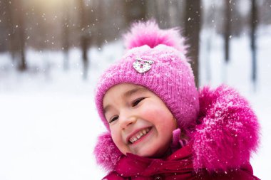 Soğukta küçük bir kız kara bakar ve gülümser. Winter, bebeği açık havada dolaştır, kar. Sıcak kıyafetler, örgü şapka, kürk ponpon ve başlık. Kartopu savaşı, kış eğlencesi. Yakın plan portre