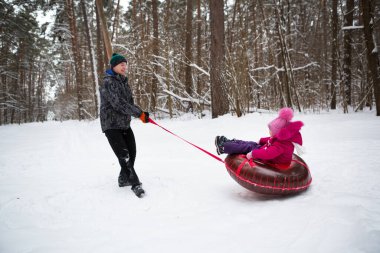 Baba, kışın kar yağarken kızına kaydırağın üstünde biner. Borular, kış sporları ve aile eğlencesi. Sıcak kıyafetler, ponponlu örülü şapka. Yardım ve destek, aile değerleri