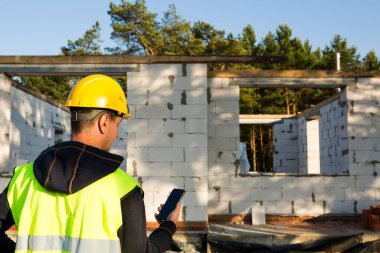 İnşaat işçisi, ev inşaatının arka planına karşı sarı baret ve yansıtıcı yelek içinde akıllı bir telefonla konuşur. Duvarlar ve pencere açıkları gözenekli beton bloklardan yapılmıştır..