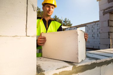 İnşaatçı elinde hücresel bir beton bloğu tutuyor. Evin duvarları. Koruyucu giysili inşaat işçileri. Sert şapka ve yansıtıcı yelek..
