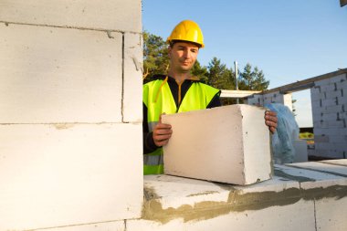 İnşaatçı elinde hücresel bir beton bloğu tutuyor. Evin duvarları. Koruyucu giysili inşaat işçileri. Sert şapka ve yansıtıcı yelek..