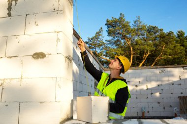 İnşaat işçisi, pencere açıklığının uzunluğunu ve duvarın uzunluğunu mezurayla ölçüyor. Kulübe gözenekli beton bloklar, koruyucu giysiler - kalın şapka ve bir yelek