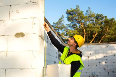 İnşaat işçisi, pencere açıklığının uzunluğunu ve duvarın uzunluğunu mezurayla ölçüyor. Kulübe gözenekli beton bloklar, koruyucu giysiler - kalın şapka ve bir yelek