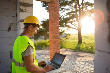 Gözenekli beton bloklardan yapılmış bir evin şantiyesinde inşaat mühendisi yansıtıcı güvenlik yeleği ve baret takan bir bilgisayarda çalışıyor. Tasarım, inşaat, çizim projesi, kontrol