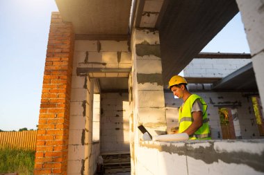 Gözenekli beton bloklardan yapılmış bir evin şantiyesinde inşaat mühendisi yansıtıcı güvenlik yeleği ve baret takan bir bilgisayarda çalışıyor. Tasarım, inşaat, çizim projesi, kontrol