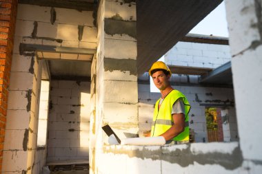 Gözenekli beton bloklardan yapılmış bir evin şantiyesinde inşaat mühendisi yansıtıcı güvenlik yeleği ve baret takan bir bilgisayarda çalışıyor. Tasarım, inşaat, çizim projesi, kontrol