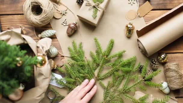 Hands pack a gift for Christmas and new year in eco-friendly materials: kraft paper, live fir branches, cones, twine, tied with a bow. Tags with mock up, natural decor, hand made, flatly. Festive mood — Stock Footage Hands Pack Gift Christmas New Year Eco Friendly Materials Kraft Royalty Free Stock Video