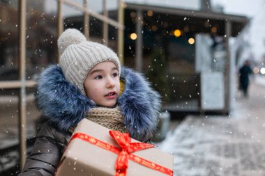 Şaşkınlık dolu bir kızın portresi, kışın bir şehir caddesinde, süsleri ve ışıkları olan karlı bir pazarda Noel için hediye kutusuyla. Sıcak kıyafetler, örgü şapka, atkı ve kürk. Boşluğu kopyala