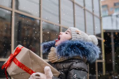 Noel için hediye kutusu olan mutlu kız kışın süsleri ve ışıkları olan karlı bir şehirde diliyle kar tanesi yakalıyor. Sıcak kıyafetler, örgü şapka, atkı ve kürk.. 