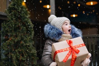 Şaşkınlık dolu bir kızın portresi, kışın bir şehir caddesinde, süsleri ve ışıkları olan karlı bir pazarda Noel için hediye kutusuyla. Sıcak kıyafetler, örgü şapka, atkı ve kürk. Boşluğu kopyala
