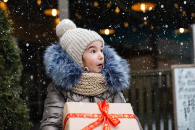 Şaşkınlık dolu bir kızın portresi, kışın bir şehir caddesinde, süsleri ve ışıkları olan karlı bir pazarda Noel için hediye kutusuyla. Sıcak kıyafetler, örgü şapka, atkı ve kürk. Boşluğu kopyala