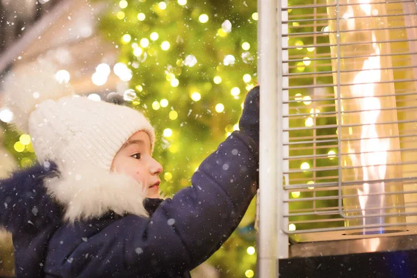 暖かい帽子の女の子は 街のクリスマスマーケットで暖まるガスバーナーの炎を見て あなたの手を暖めます クリスマスツリー お祝い事に焦点を当てたガーランドのライト ストック画像