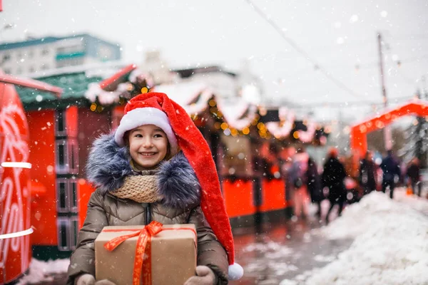 冬に街の路上でクリスマスのためのギフトボックスとサンタの帽子の喜びの女の子の肖像画は 装飾や妖精の光とお祝いの市場で雪と 暖かい服 ニットスカーフと毛皮 ストックフォト