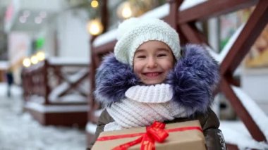 Noel hediyesi kutulu neşeli bir kızın portresi kışın bir şehir caddesinde süsleri ve peri ışıklarıyla karlı bir pazarda. Sıcak kıyafetler, örgü şapka, atkı ve kürk. Yeni Yıl