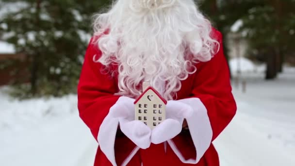 雪の中でサンタクロースの屋外の手の中にミニチュアコテージ 不動産 住宅ローンを扱う 居心地の良い家 贈り物の配達 メリークリスマス新年の予約 — ストック動画