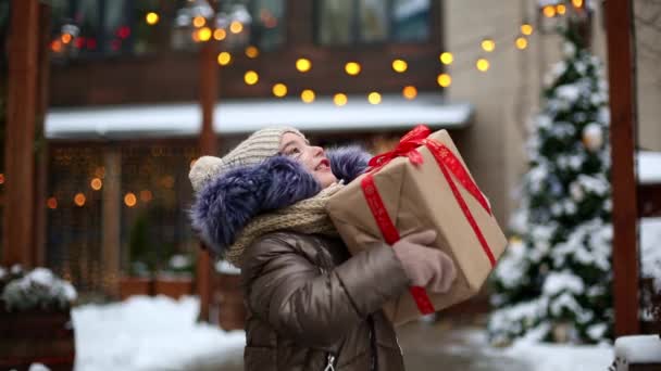 冬の街の路上でクリスマスのためのギフトボックスを持つ喜びの少女の肖像画 装飾や妖精の光を持つお祝い市場で雪 暖かい服 ニット帽 スカーフ — ストック動画
