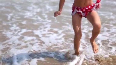 A little girl in a red swimsuit is playing on the beach with a sea wave - jumping, running, having fun. Swimming, traveling, playing with water