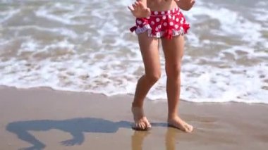 A little girl in a red swimsuit is playing on the beach with a sea wave - jumping, running, having fun. Swimming, traveling, playing with water