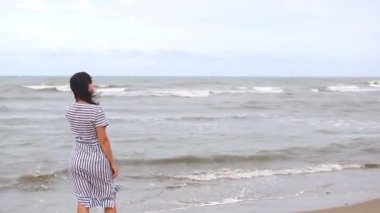 A brunette woman in a striped dress with long hair fluttering in the wind stands by the sea, looks at the waves, holds and removes her hair from her face. Tourism, beach holidays, breeze