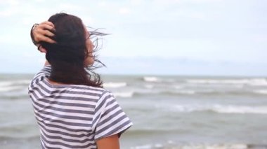 A brunette woman in a striped dress with long hair fluttering in the wind stands by the sea, looks at the waves, holds and removes her hair from her face. Tourism, beach holidays, breeze