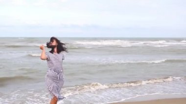 A brunette woman in a striped dress with long hair fluttering in the wind stands by the sea, looks at the waves, holds and removes her hair from her face. Tourism, beach holidays, breeze