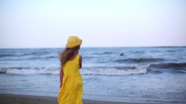 身穿黄色连衣裙 头戴黄帽 长长的头发在微风中飘扬的女孩望着大海 摇曳着 背对着镜框站在那里 假期结束 告别大海 — 图库视频影像