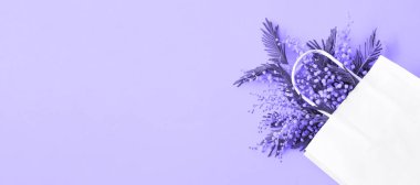 A bouquet of mimosa in a white mock up gift bag. Spring shopping, gifts and promotions for International Women's Day. Purple background, very peri tinting, copyspace, banner