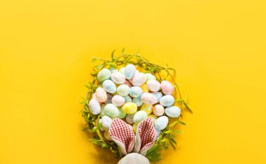 Genç bahar yapraklarından oluşan bir çelenk renkli Paskalya yumurtaları ve paskalya tavşanı kulaklarıyla dallara konuyor. Sarı arka planda yumurtalı yuva.