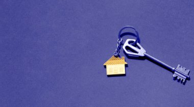 Key chain in the shape of wooden house with key on a purple background. Very peri tinting. Building, design, project, moving to new home, mortgage, rent and purchase real estate. Copy space