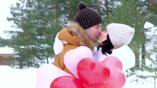 愛のキスで男と女性と心の形でピンクと赤い風船の贈り物と雪の中で冬の屋外の日付に抱擁 バレンタインデー 散歩中の愛の幸せなカップル スローモーション — ストック動画