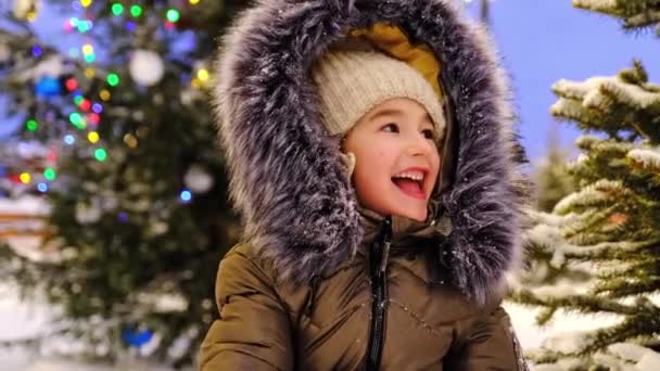 妖精の光の背景に毛皮のフードで冬の幸せな女の子の肖像画 冬の屋外で夜に雪の中でクリスマスツリー 雪を投げ 彼女の手を拍手 笑いを再生します クリスマス — ストック動画