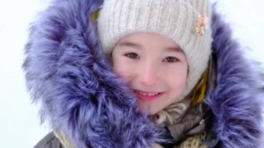 Kış sıcacık giysileri içinde neşeli ve mutlu 5 yaşında bir kızın portresi. Örgü şapka, kürklü başlık, ceket. Beyaz karın arka planına karşı. Yavaş Hareket