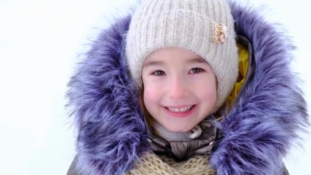 冬の暖かい服を着た明るく幸せな5歳の少女の肖像画 ニット帽 毛皮のフード ジャケット 白い雪を背景に スローモーション — ストック動画
