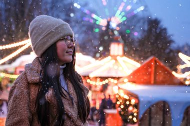 Girl on background of carousels and amusement park in winter. Woman looks at night Christmas city.