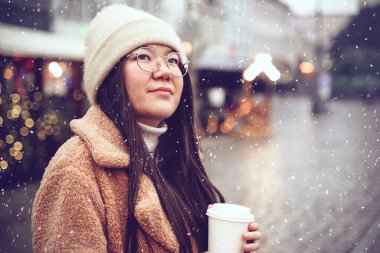 Asian woman enjoy coffee and snowfall outdoors. Cute girl in snowy winter weather