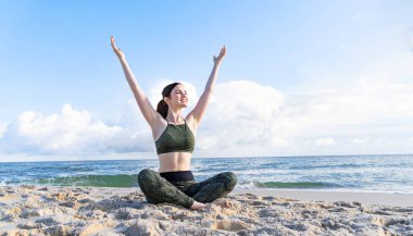 Happy woman enjoying life and nature with hands up. Healthy and harmonious pretty girl sitting by sea against blue sky