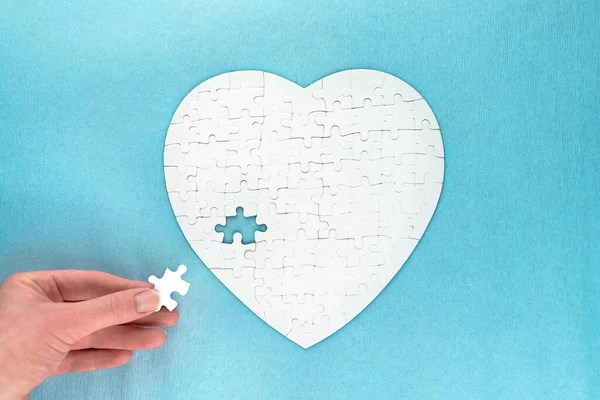 White puzzles in shape of heart. Woman\'s hand collects jigsaw on blue background