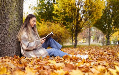 Pretty girl reads book in autumn park. Young woman sits on background of nature. Rest pastime leisure