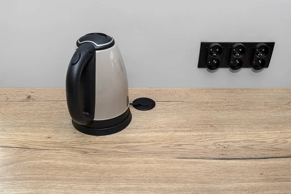 An electric kettle standing on the countertop on the kitchen cabinets, visible hole in the table top through which the electric cable goes.