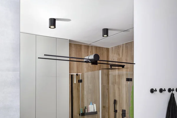 Black sconce mounted in a large mirror in the bathroom, visible LED lighting in the shape of a tube and shower cubicle reflected in a mirror.