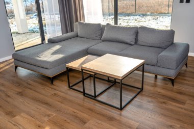 Gri bir kanepesi olan modern ve basit bir oturma odası, görünen büyük teras pencereleri ve biri diğerinin altında iki masa..